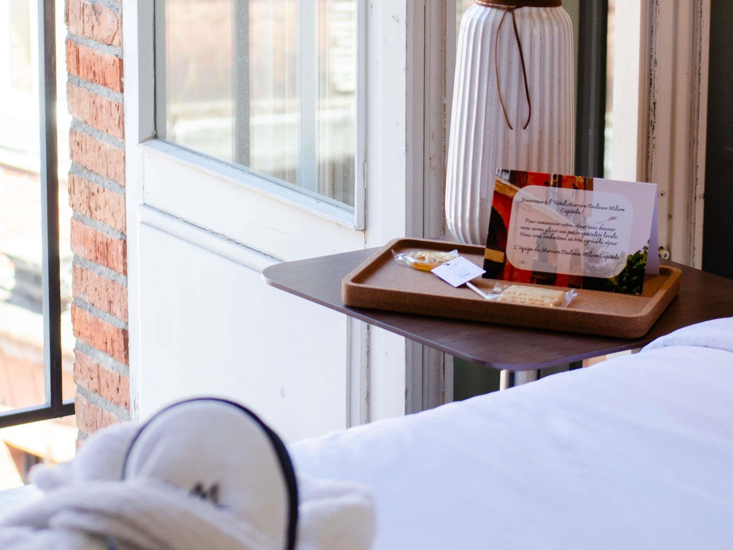 A hotel room with a bed, a window with a view of the outside, and a tray with a welcome card and some food items.