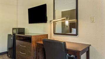 A compact hotel room with a wooden desk, a black chair, a flat-screen TV mounted on the wall, and a microwave on the left.