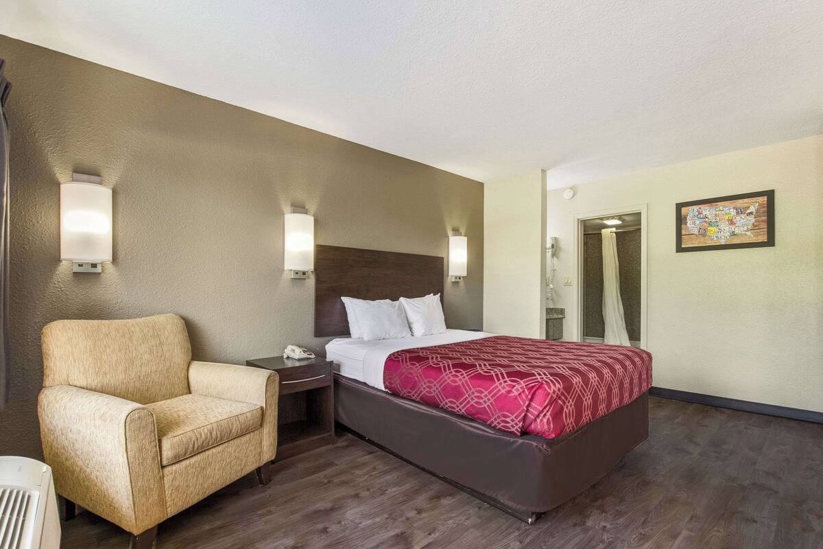 A hotel room with a bed, a chair, a nightstand, a phone, and a wall-mounted light fixture.