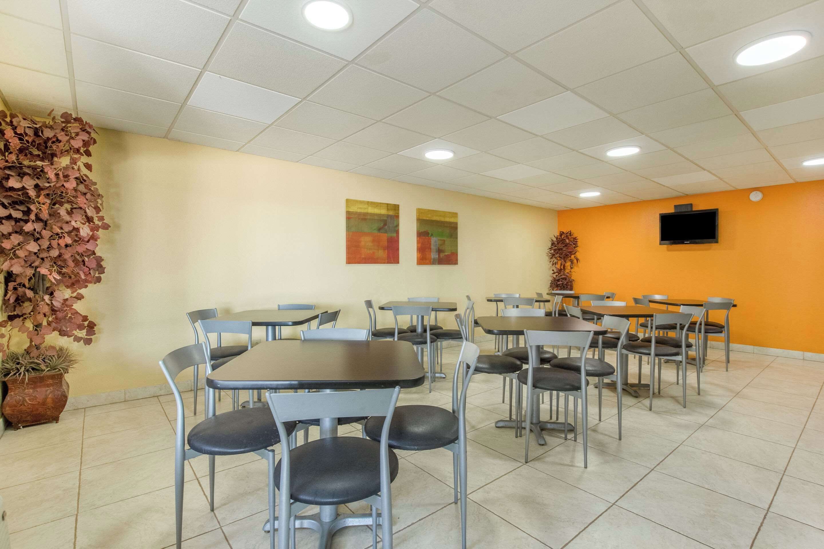 Un espace repas avec plusieurs tables et chaises, un téléviseur fixé au mur et une plante en pot sur la gauche.