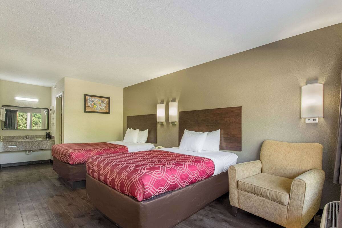 A hotel room with two beds, a chair, a bathroom with a sink and mirror, and a painting on the wall.