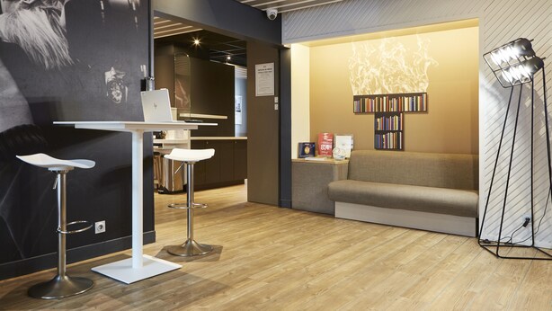 A modern interior with a wooden floor, a white table with a laptop, bar stools, a sofa, and a bookshelf.