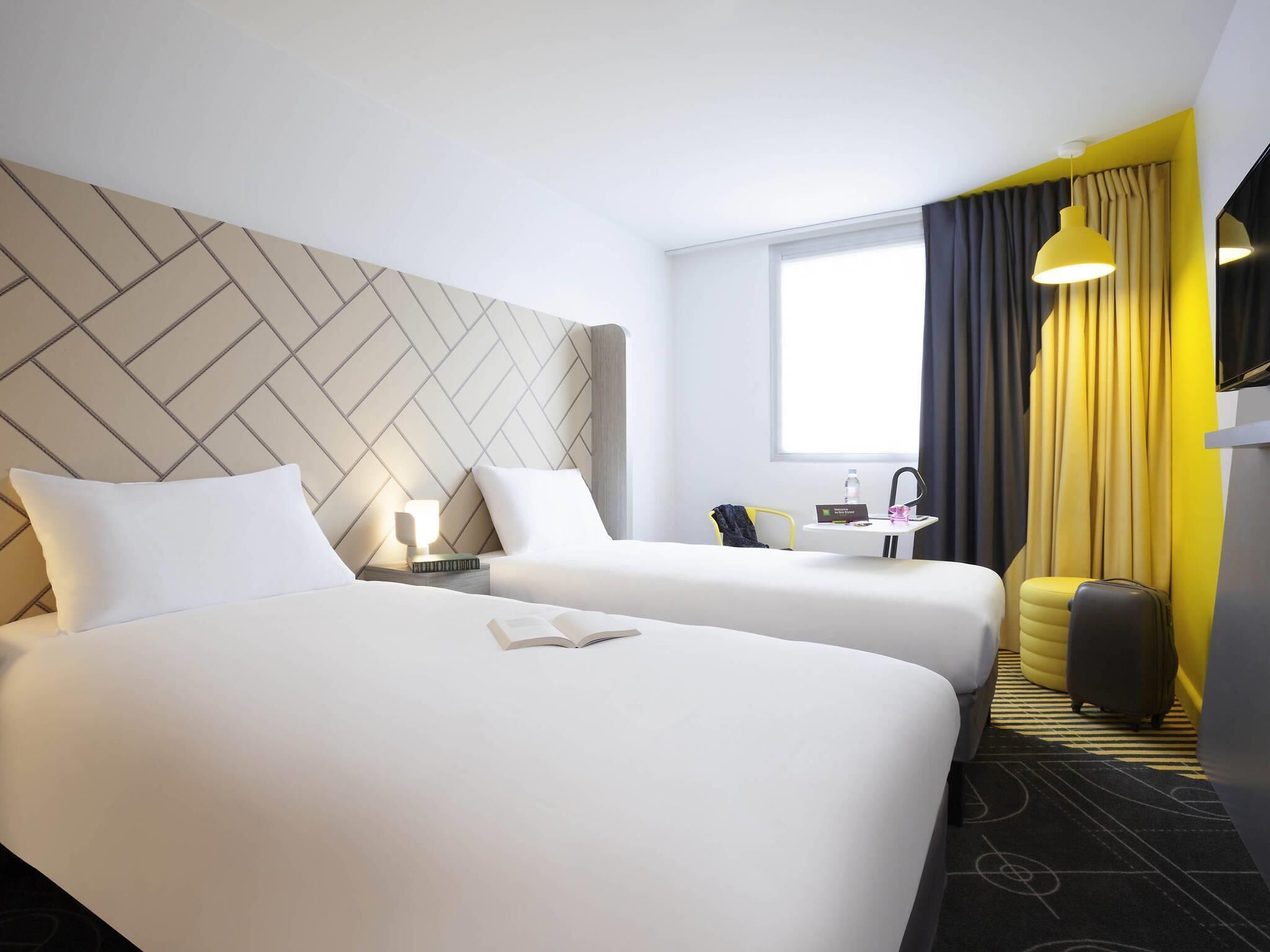 A hotel room with two beds, a desk, a chair, a suitcase, and a window with curtains.