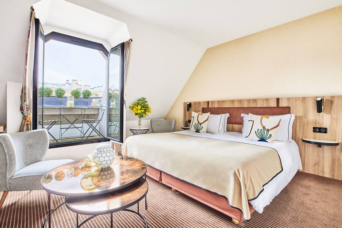 A hotel room with a large bed, a round table with a vase, a chair, and a view of buildings through a large window.