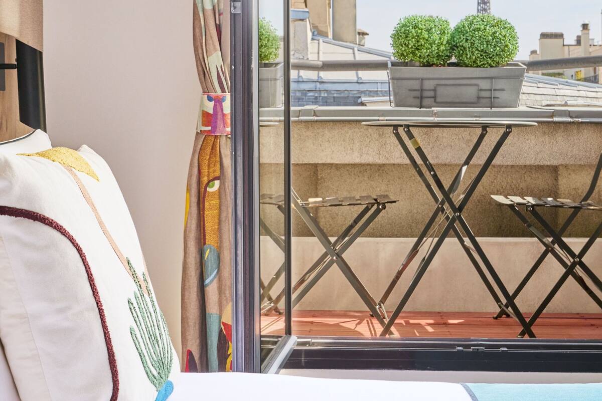 A room with a bed, a window, and a view of buildings and a tower.