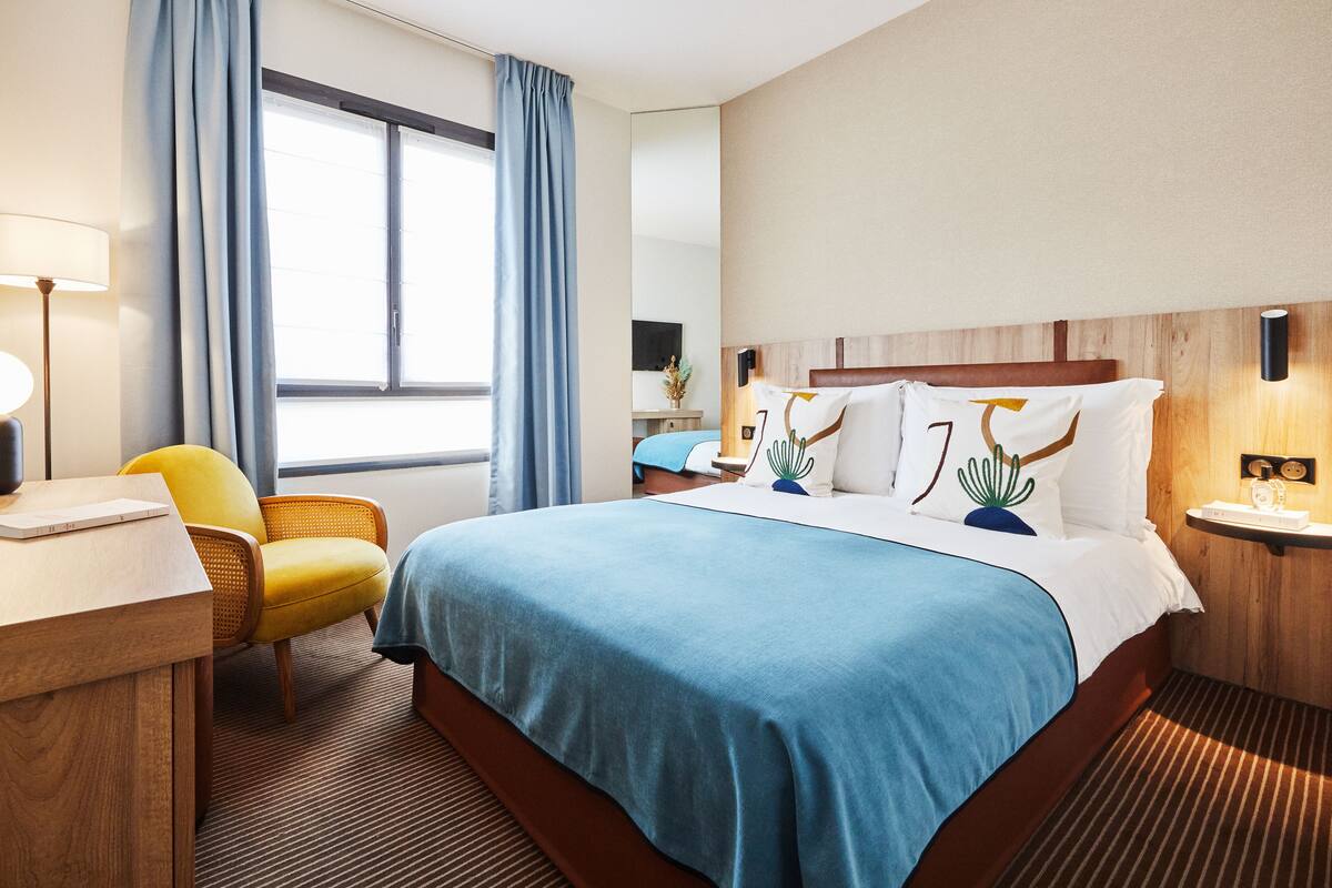 A hotel room with a large bed, a desk with a lamp, a chair, and a window with curtains.