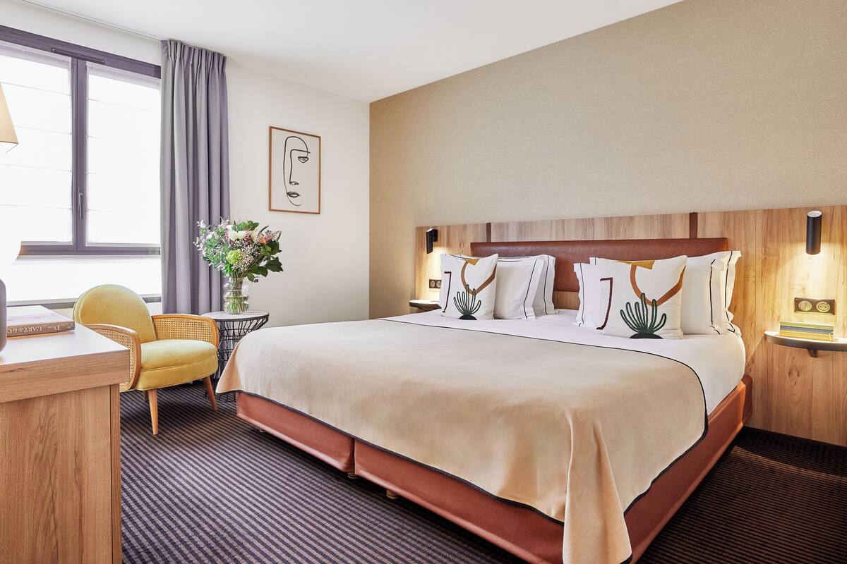 A hotel room with a large bed, a desk, a chair, a window with curtains, a wall-mounted lamp, and a framed artwork.