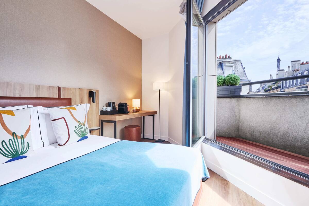 A hotel room with a large bed, a desk with a lamp, and a view of the cityscape.