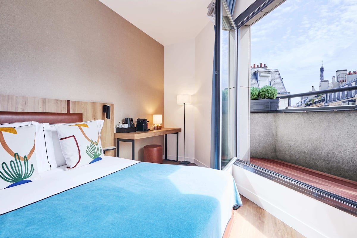 A hotel room with a large bed, a desk with a lamp, and a view of the cityscape.