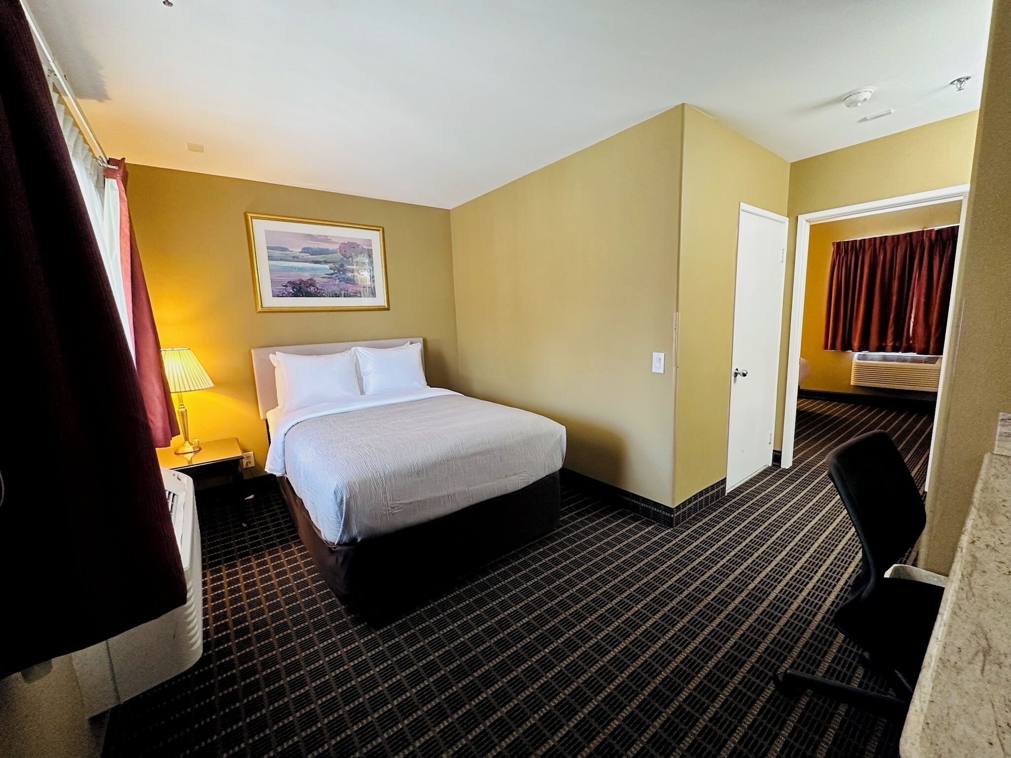 A hotel room with a bed, a nightstand, a lamp, a painting on the wall, and a door leading to another room.