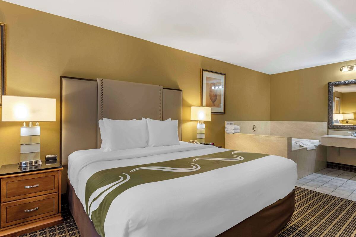 A hotel room with a large bed, bedside lamps, a nightstand, a bathroom with a sink and mirror, and a framed picture on the wall.