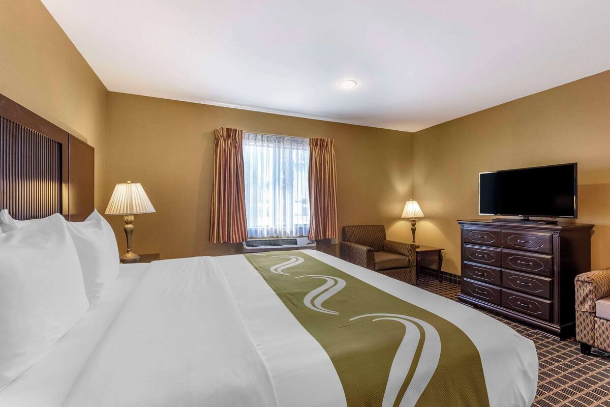 A hotel room with a large bed, a television, a dresser, and a window with curtains.