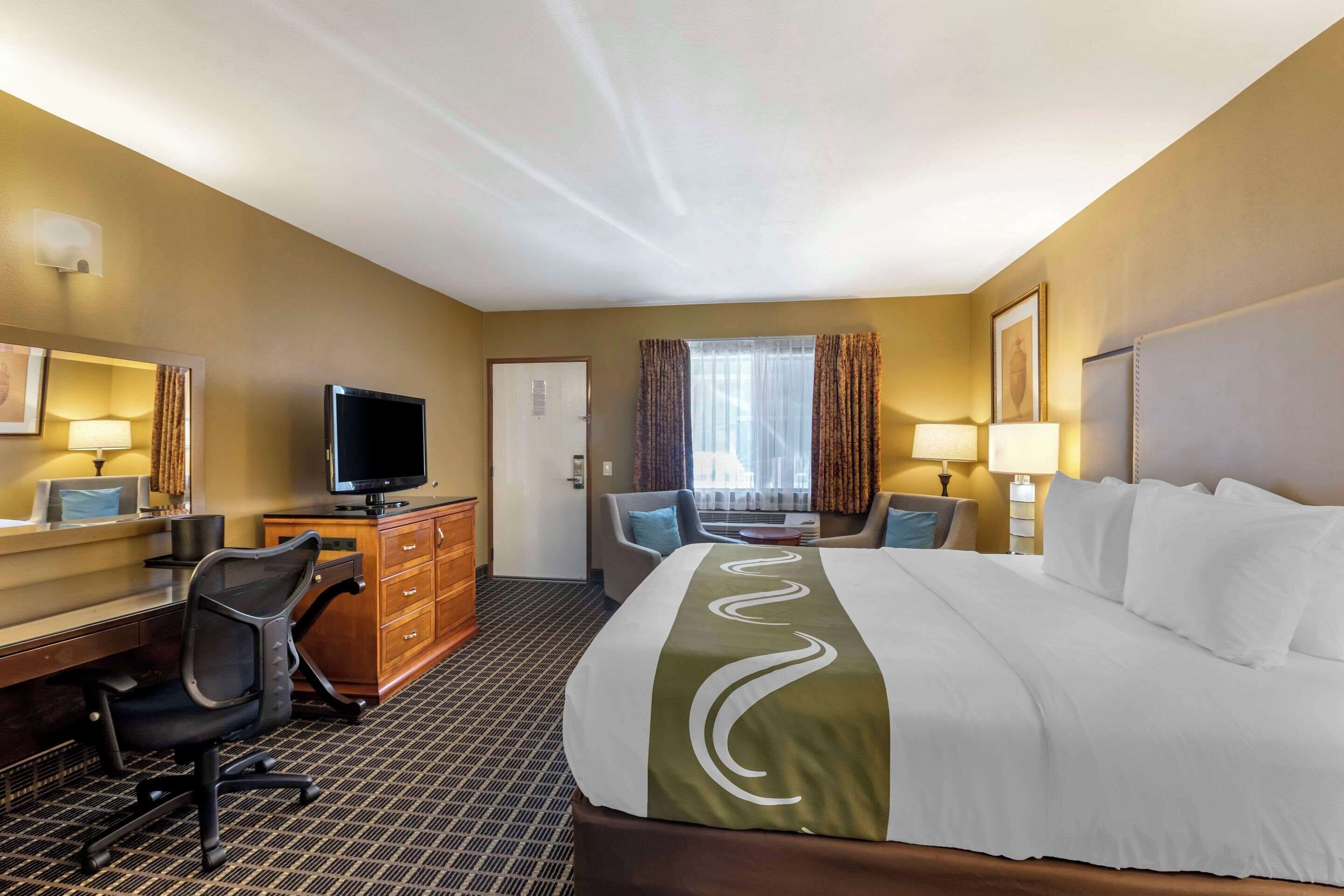 A hotel room with a large bed, a desk with a computer, a chair, a television, and a window with curtains.