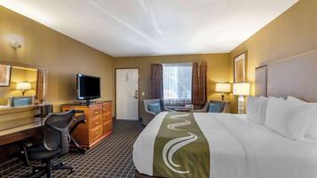 A hotel room with a large bed, a desk with a computer, a chair, a television, and a window with curtains.