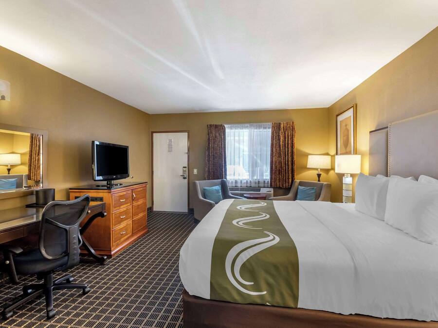 A hotel room with a large bed, a desk with a computer, a chair, a television, and a window with curtains.