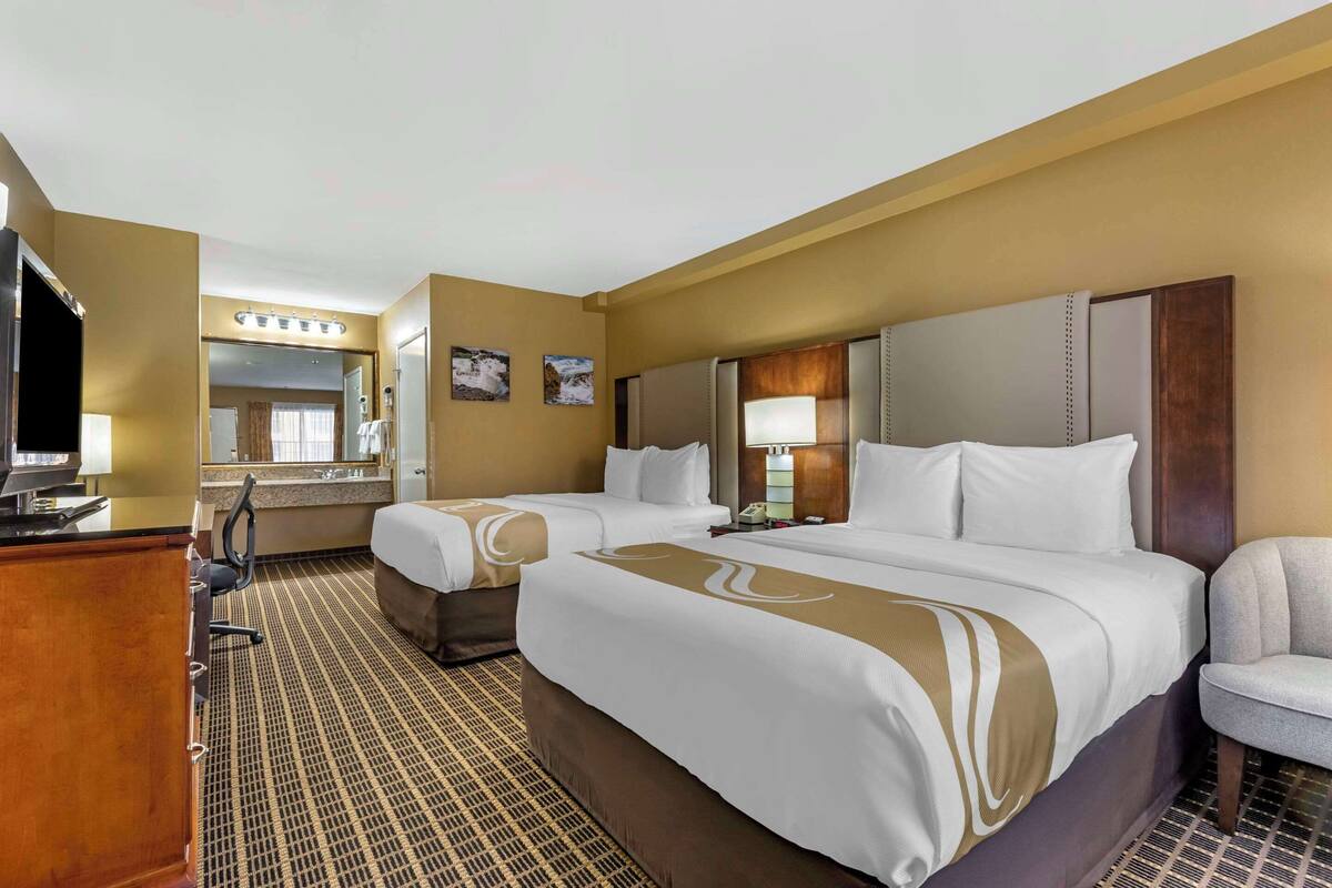 A hotel room with two beds, a desk, a chair, a TV, and a bathroom visible through an open door.