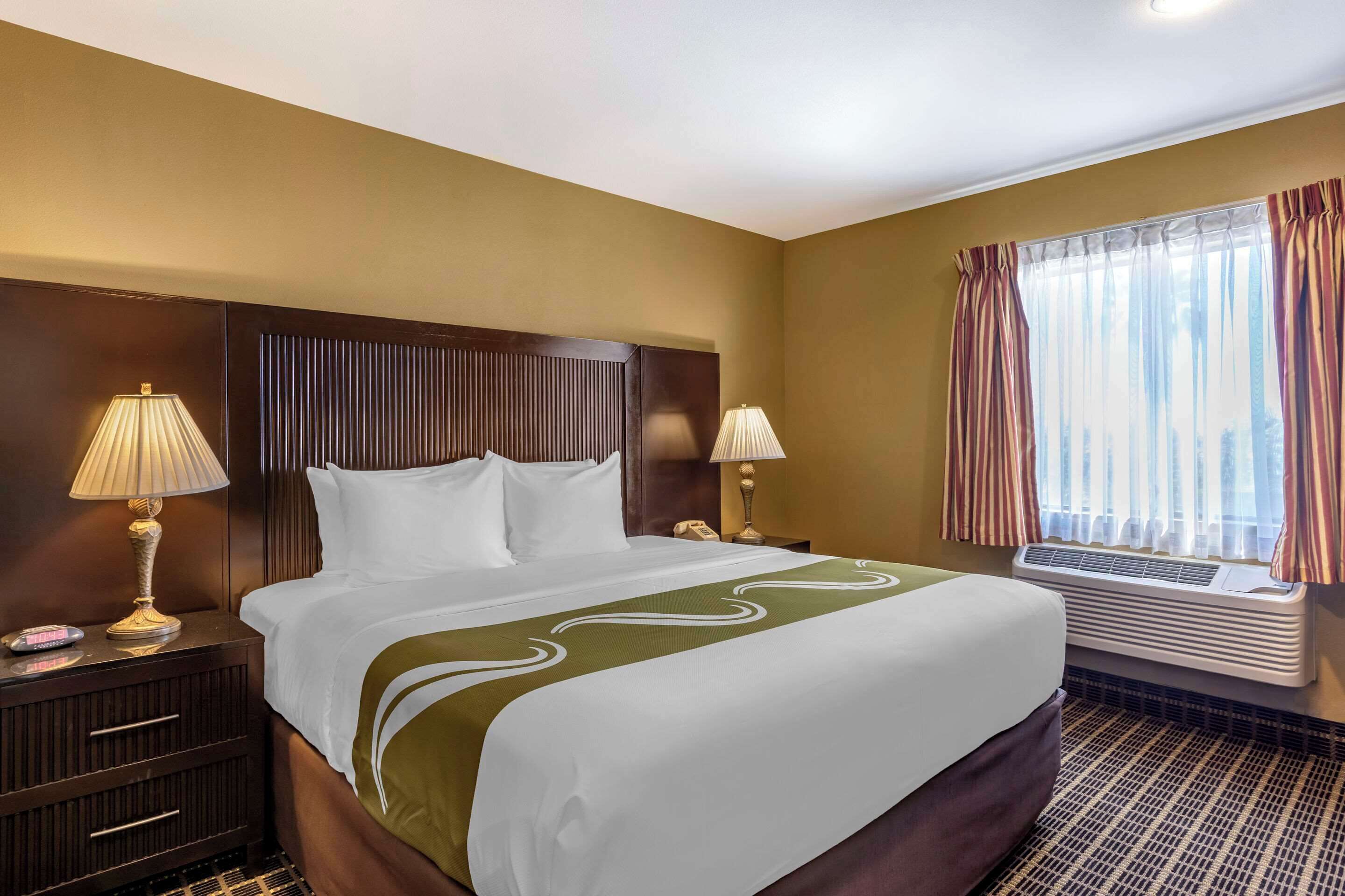 A hotel room with a large bed, two bedside lamps, a telephone, an air conditioning unit, and a window with curtains.