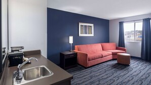 Habitación de hotel con kitchenette, un sofá, un pequeño reposapiés, un cuadro enmarcado en la pared y una ventana con cortinas.