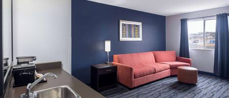 A hotel room with a kitchenette, a sofa, a small ottoman, a framed picture on the wall, and a window with curtains.