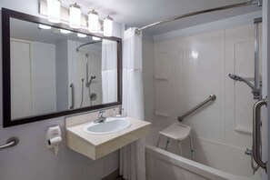 A modern bathroom with a white sink, a large mirror, a handheld showerhead, and a grab bar. - Travelodge Suites by Wyndham Saint John (Saint John)