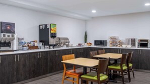 Una cocina moderna con cafetera, refrigerador y microondas. Hay mesas y sillas de madera con cojines verdes.
