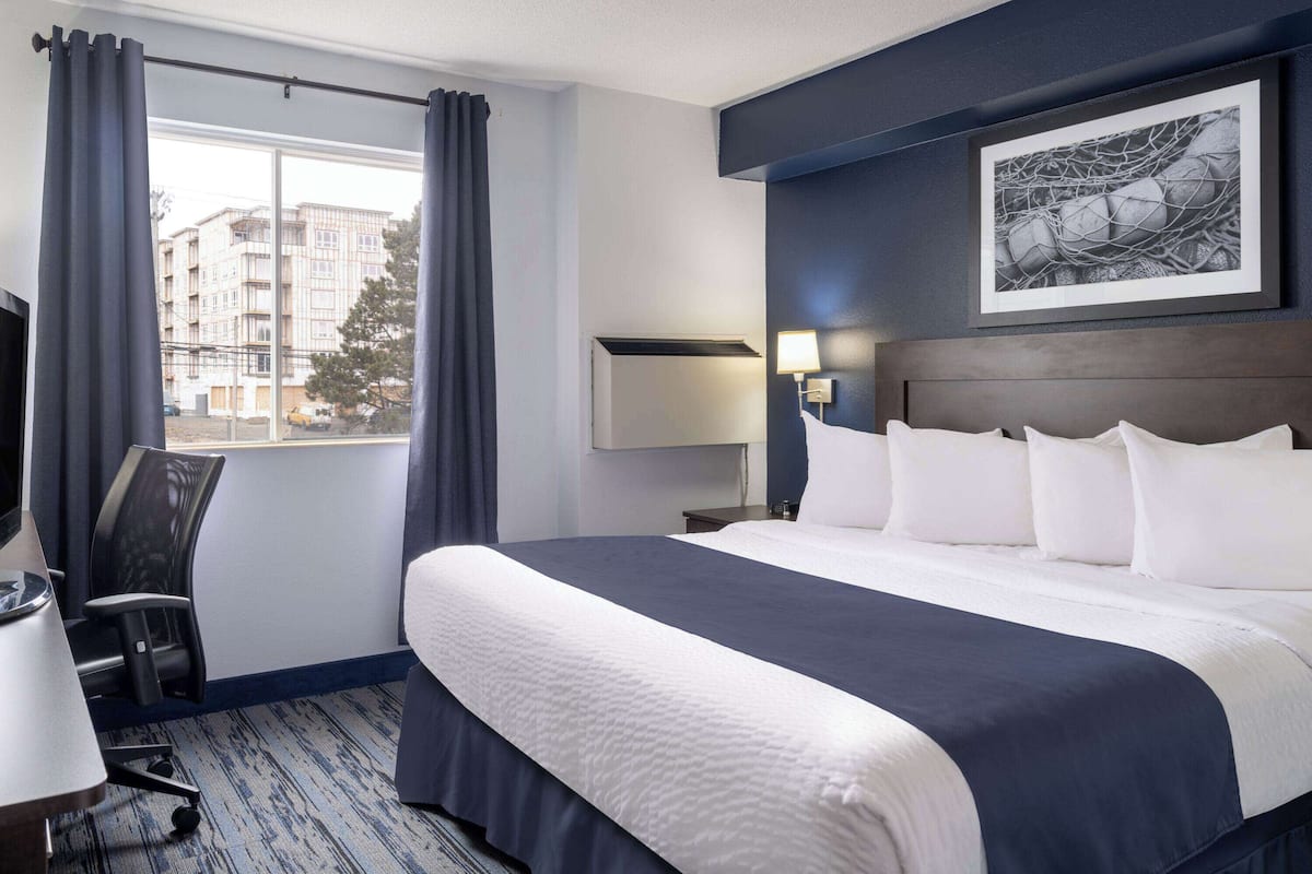 A hotel room with a large bed, a desk, a chair, a window with curtains, a wall-mounted lamp, and a framed picture.
