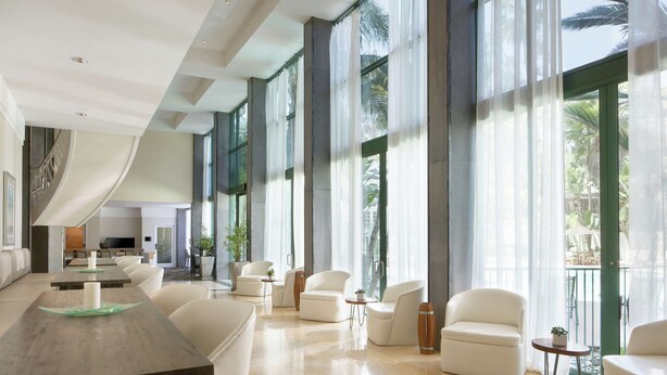 A spacious hotel lobby with a long reception desk, multiple seating areas, and large windows offering views of the outdoors.