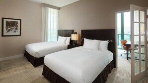 A hotel room with two beds, a nightstand with a lamp, a framed picture on the wall, and a view through an open door.