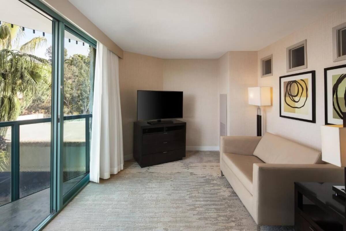 Et moderne hotelværelse med en stor skydedør, der fører til en balkon, et fladskærms-TV på et mørkt træstel, en beige sofa og en lampe monteret på væggen.