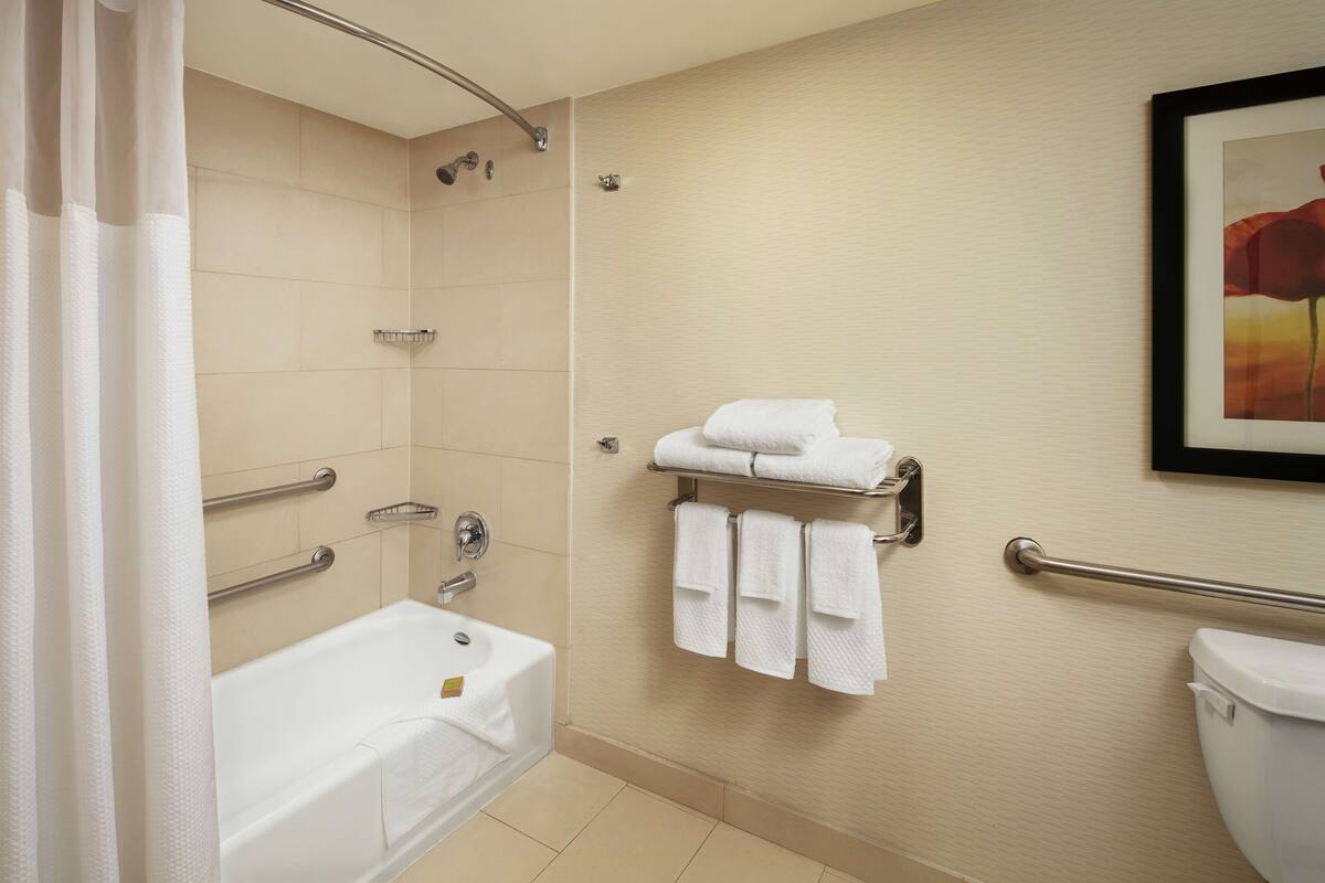 Et badeværelse med badekar, bruser og toilet. Der er gelændere og et håndklædeholder med håndklæder.