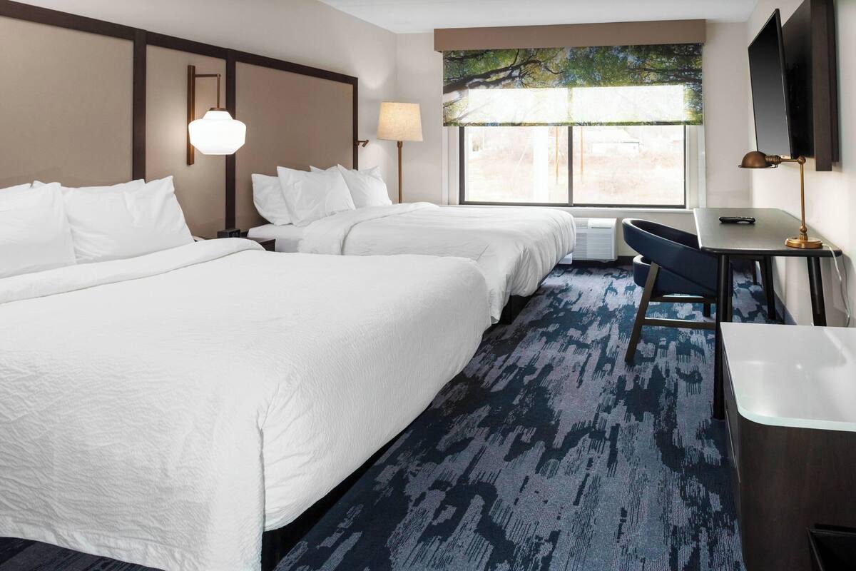 A hotel room with two beds, a desk, a chair, a TV, and a window with a view.