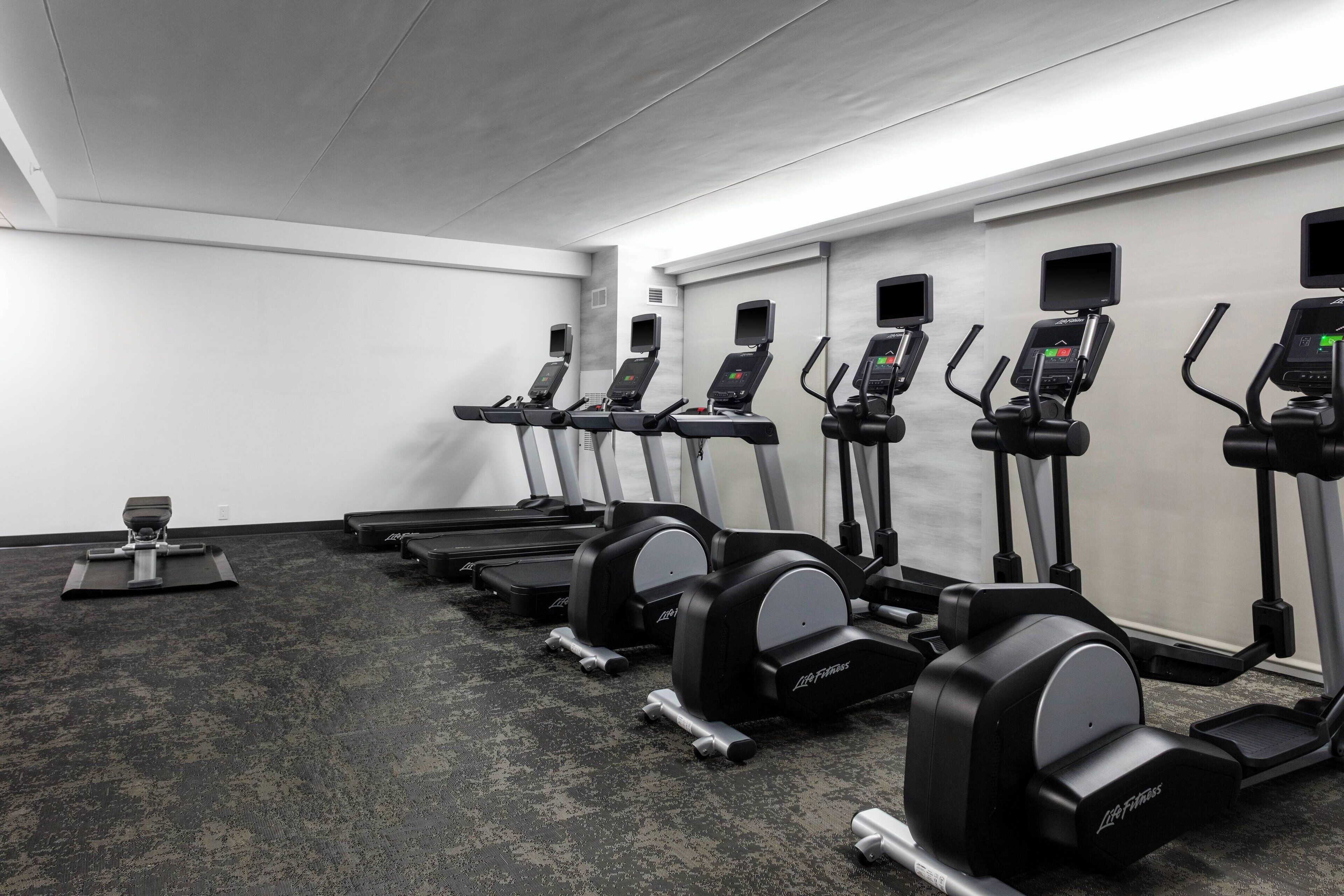 A row of cardio machines in a gym with a grey carpet and white walls.