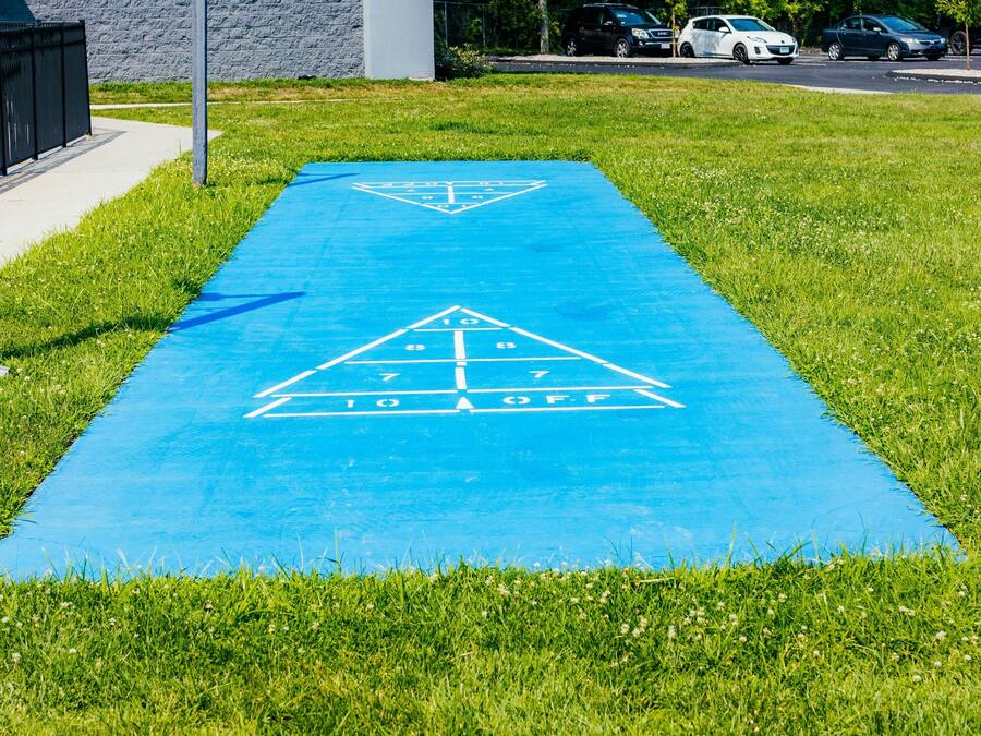 A blue hopscotch court on a grassy area with a building and parked cars in the background.
