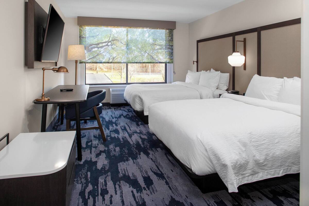 A hotel room with two beds, a desk with a chair, a TV mounted on the wall, and a large window with a view of trees.