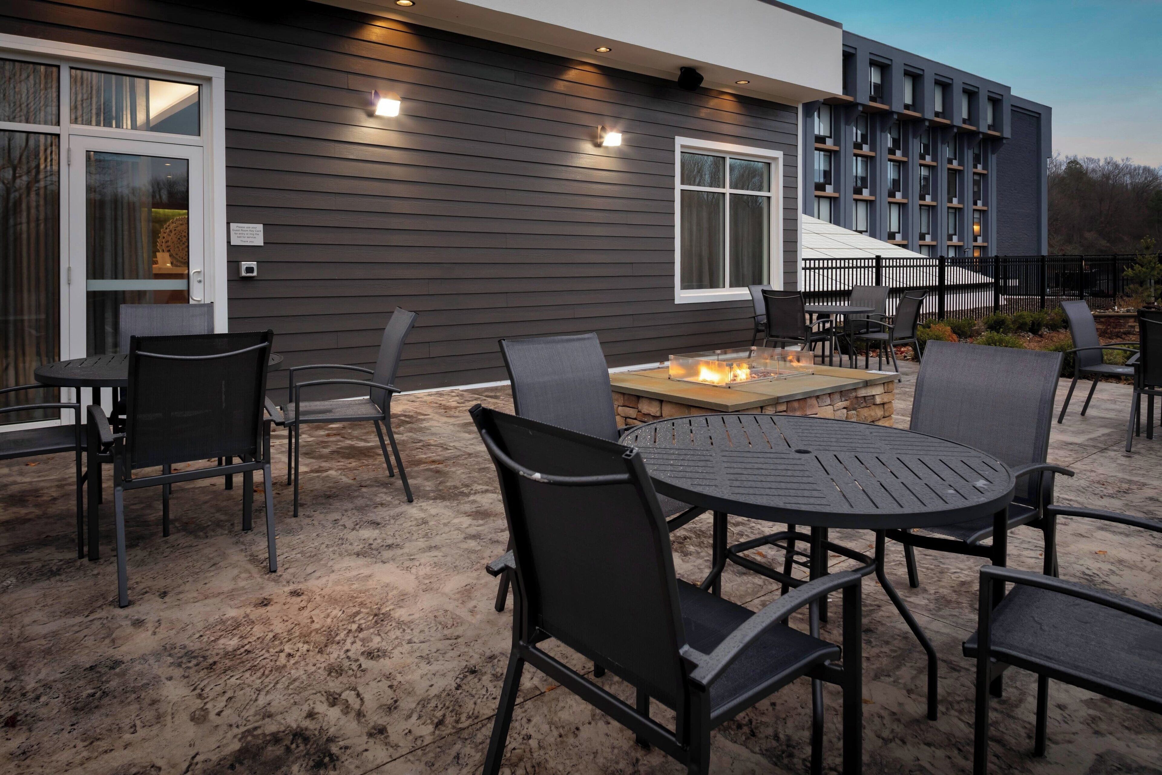 Un patio al aire libre con una mesa redonda y sillas, un hogar de fuego y un edificio con revestimiento oscuro y ventanas amplias.