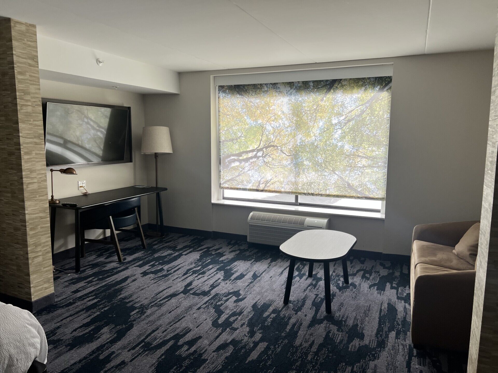 Una habitación de hotel con un ventanal grande, un escritorio con lámpara, una silla y un sofá.