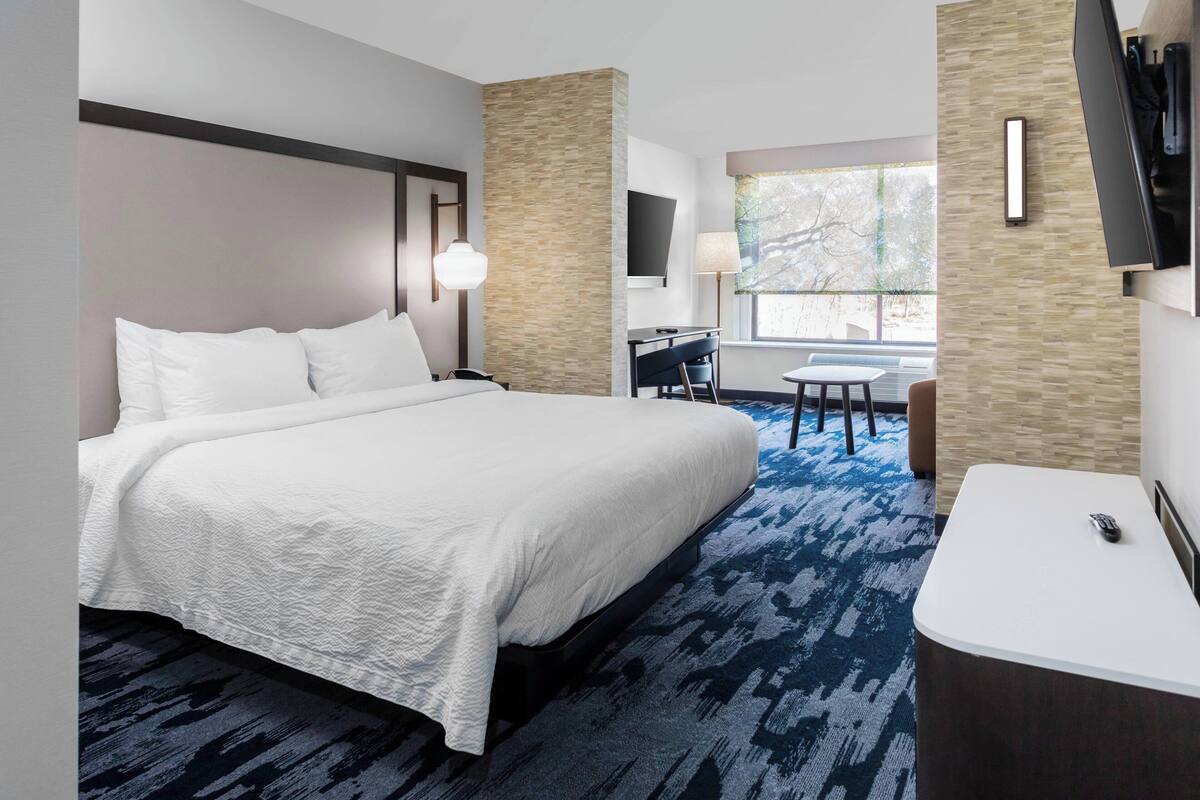 A hotel room with a large bed, a desk, a chair, a TV, and a window with a view of trees.
