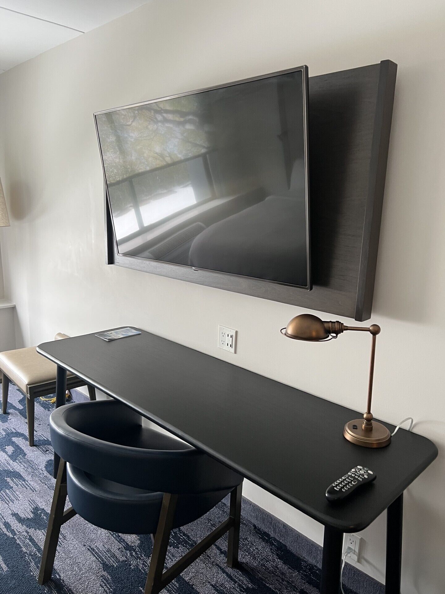 A modern hotel room with a large flat-screen TV mounted on the wall, a desk with a chair, a lamp, and a remote control on the desk.