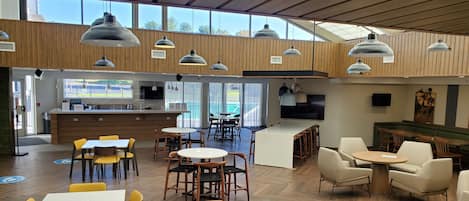 Un espacio interior amplio con diversas opciones de asientos, incluyendo mesas y sillas, una isla central con taburetes y una cocina con mostrador y electrodomésticos.