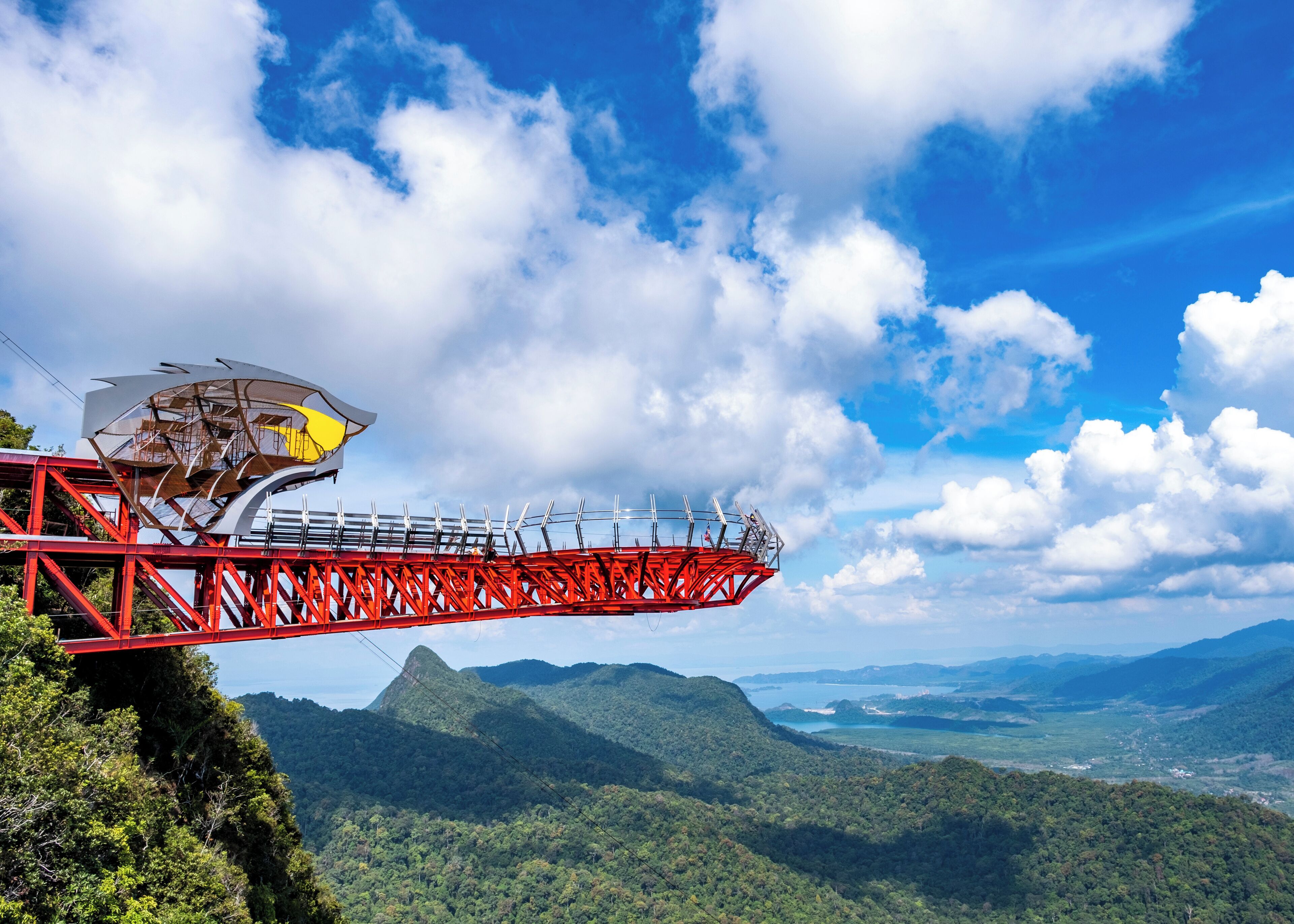 A red cable car system with passengers, set against a backdrop of lush green mountains and a clear blue sky.