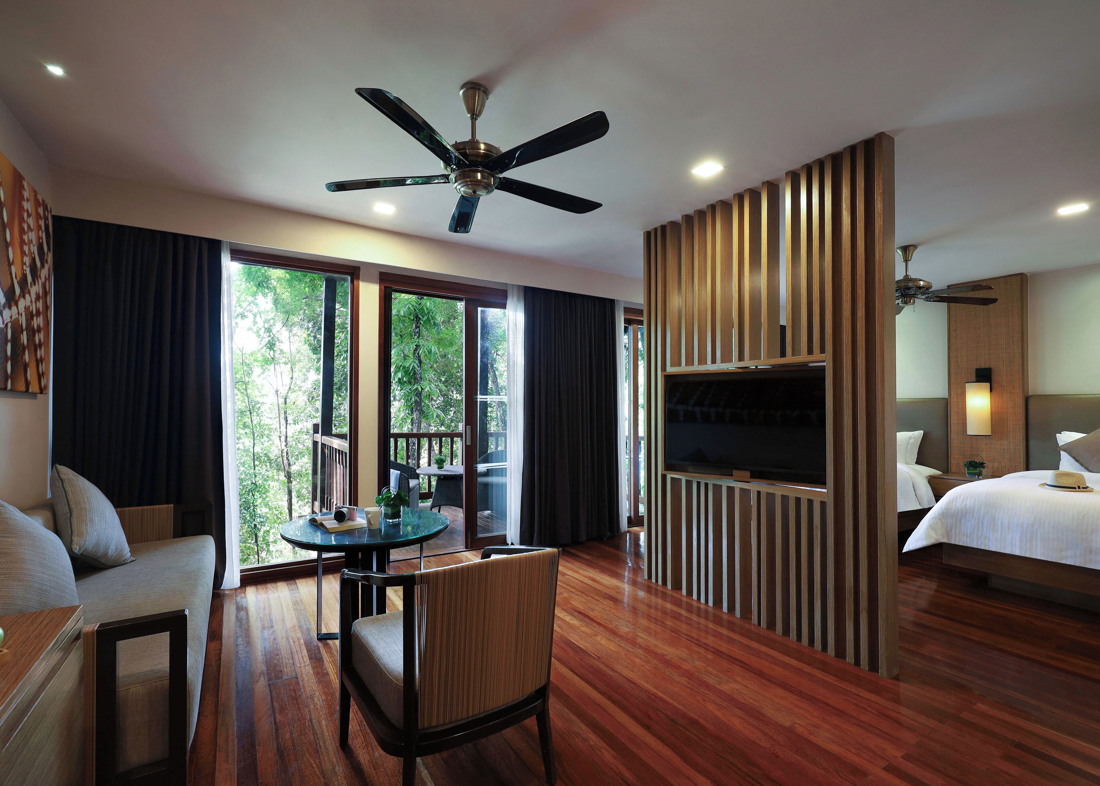 Une chambre d’hôtel avec un plancher en bois, un ventilateur de plafond, une télévision, un lit, un bureau et une fenêtre donnant sur des arbres.