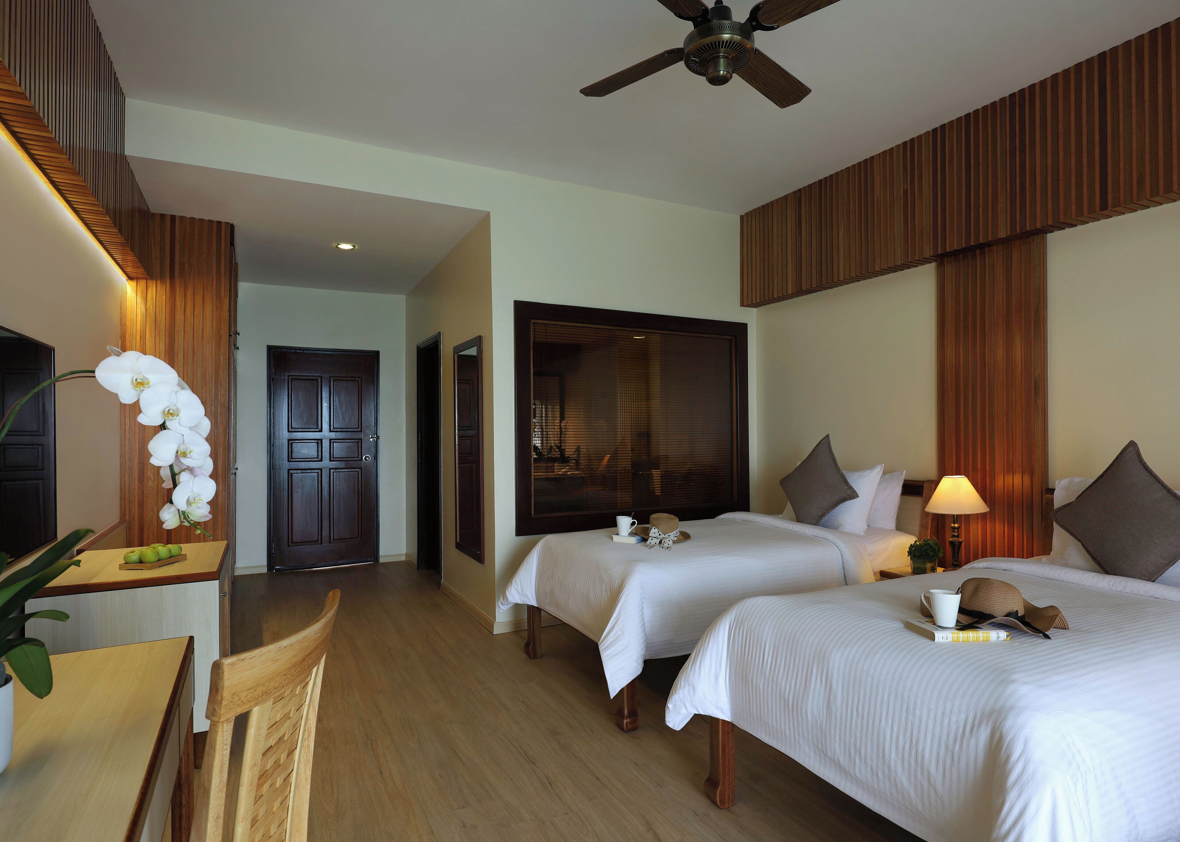 A hotel room with two beds, a desk with a chair, a ceiling fan, and a large window with blinds.