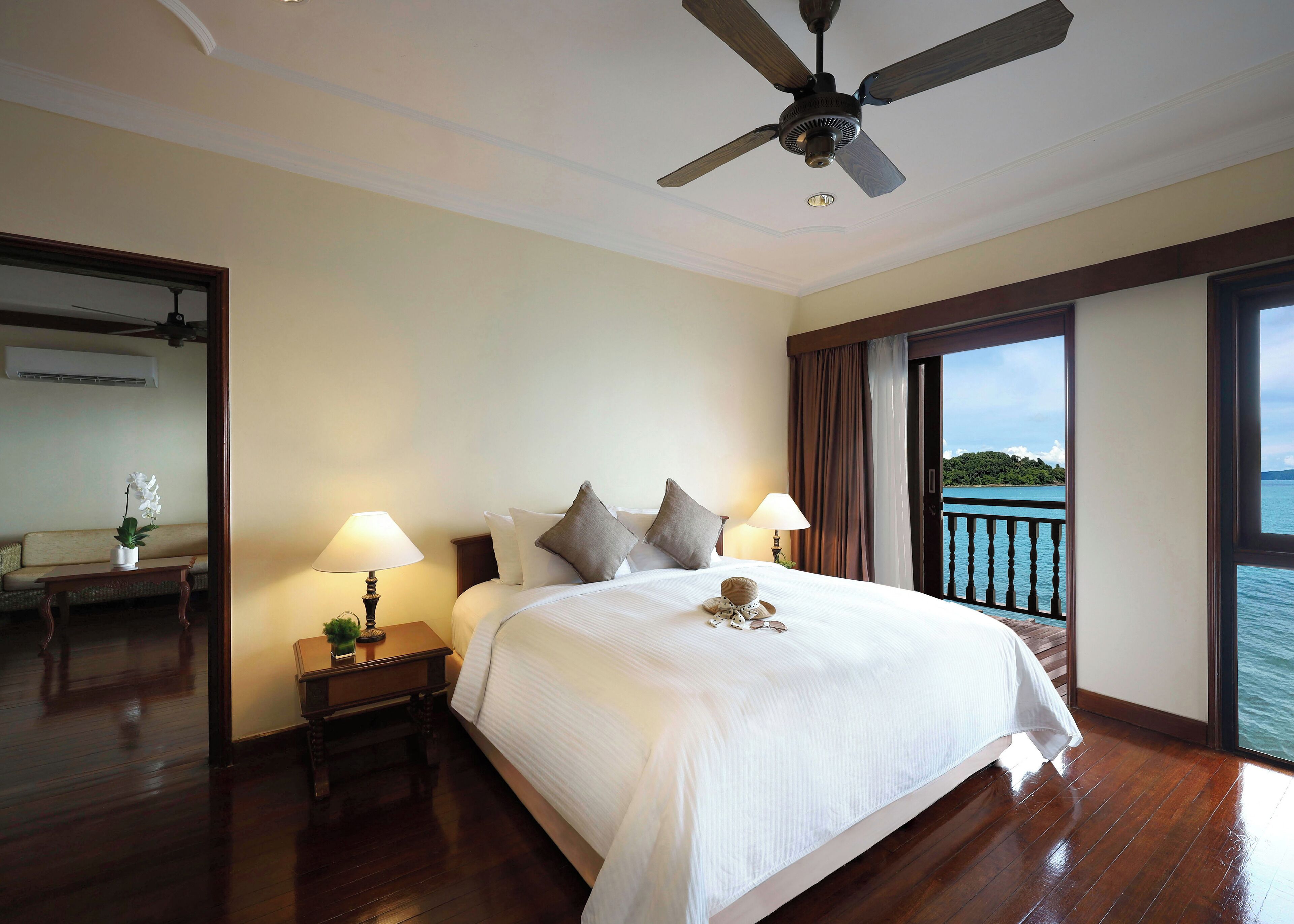 Une chambre d’hôtel avec un grand lit, deux tables de chevet avec des lampes, un ventilateur de plafond et une vue sur la mer par une grande fenêtre.