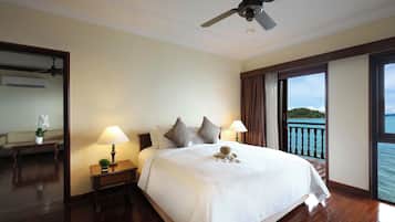 Une chambre d’hôtel avec un grand lit, deux tables de chevet avec des lampes, un ventilateur de plafond et une vue sur la mer par une grande fenêtre.