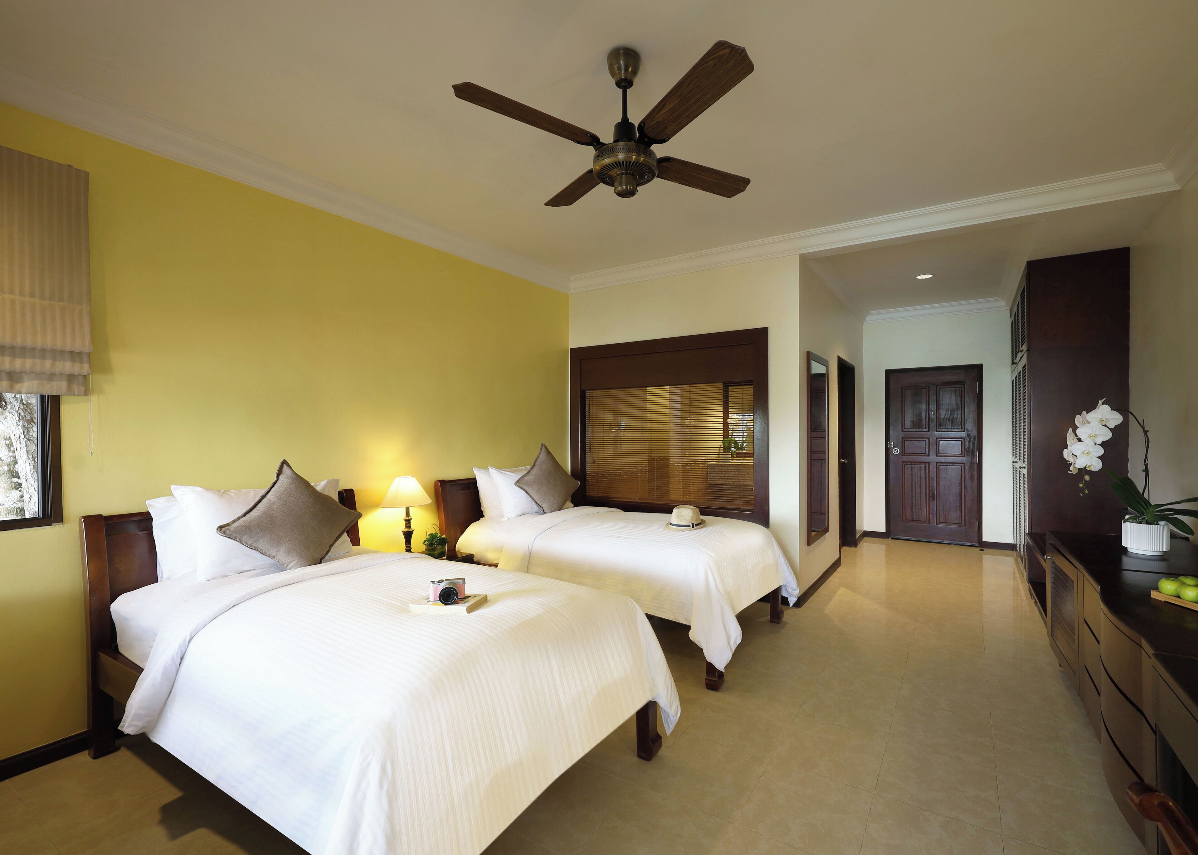Una habitación de hotel con dos camas, ventilador de techo, mesita de noche con lámpara y cabecera de madera.