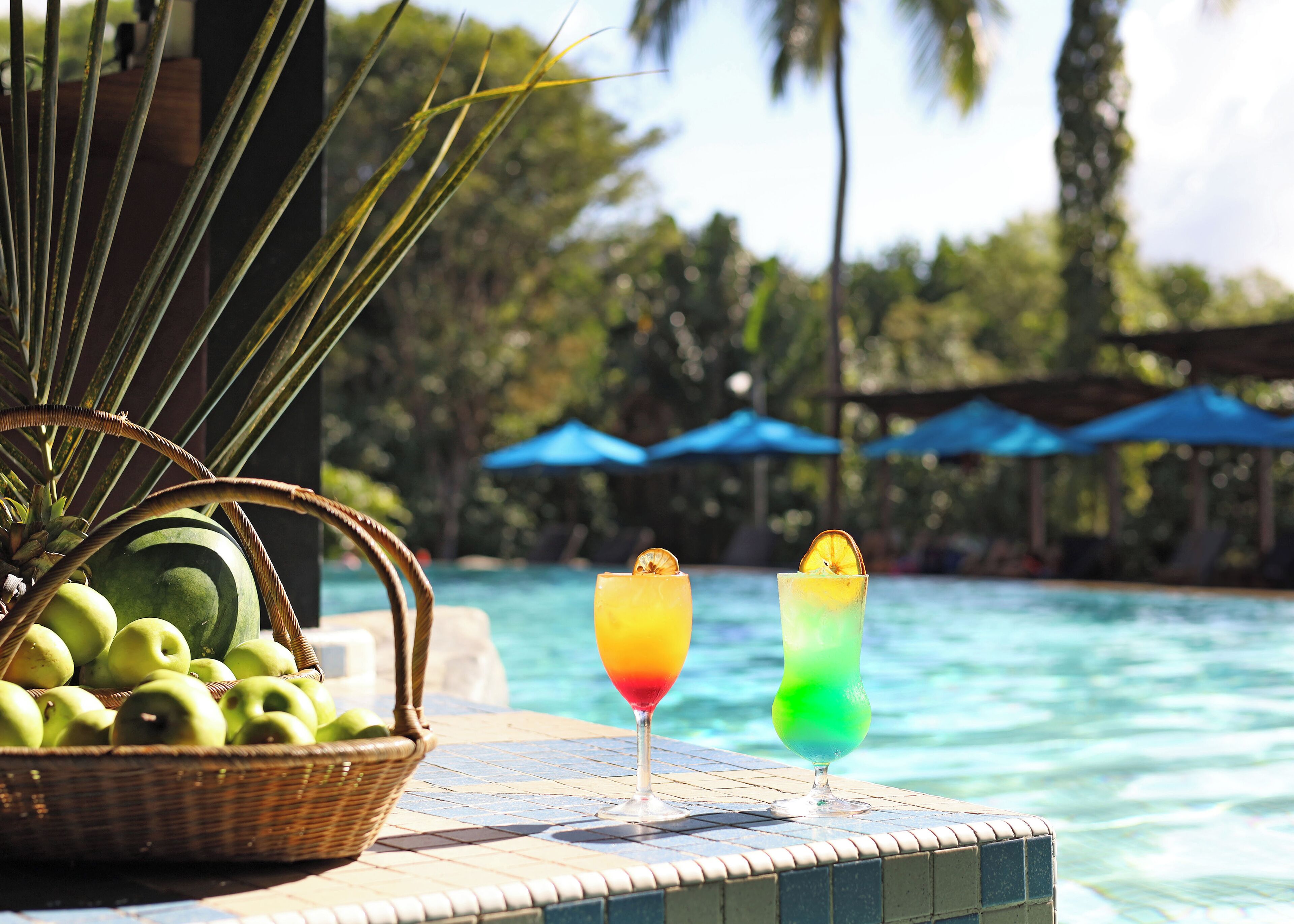 Ein Poolbereich mit einem Korbwicker-Tablett voller grüner Äpfel, zwei farbenfrohe Getränke mit Garnitur, und im Hintergrund ein freier Blick auf Pool und Palmen.