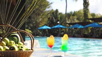 A poolside setting with a wicker basket of green apples, two colorful drinks with garnishes, and a clear view of the pool and palm trees in the background.