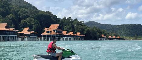 A person on a jet ski in clear water with a backdrop of overwater bungalows and lush green hills.