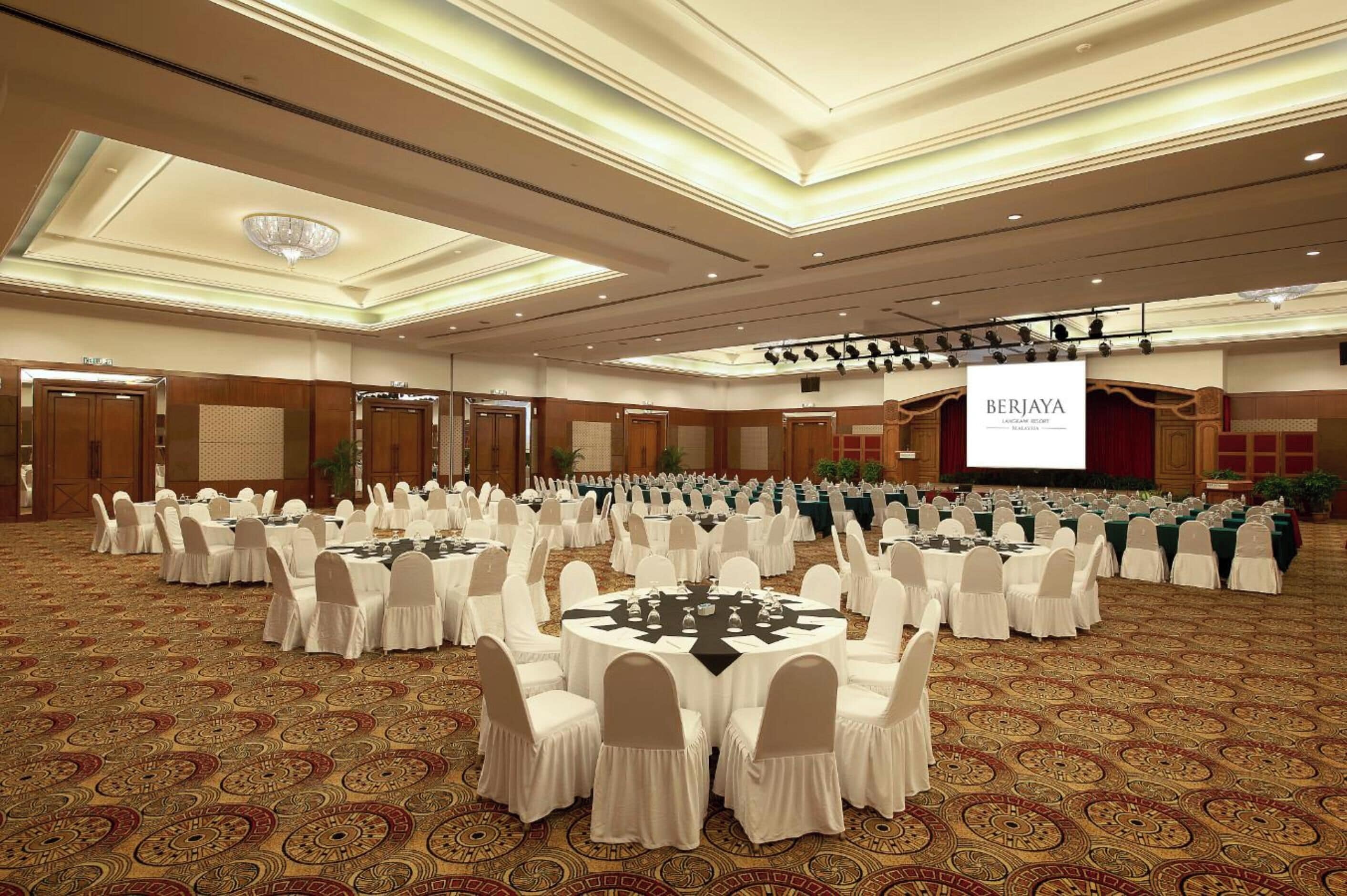 A large conference room with round tables and white chairs, a large screen, and a chandelier.