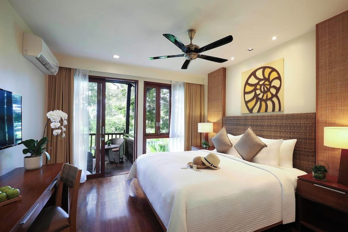 A hotel room with a bed, a desk with a chair, a ceiling fan, and a view of the outdoors.